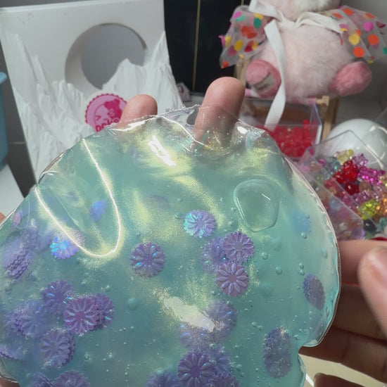 Shimmery mermaid‑blue transparent squishy pouch decorated with holographic purple daisies. Ultra‑soft texture, flows smoothly with only a light squeeze, calming aesthetic viral stress relief fidget toy.